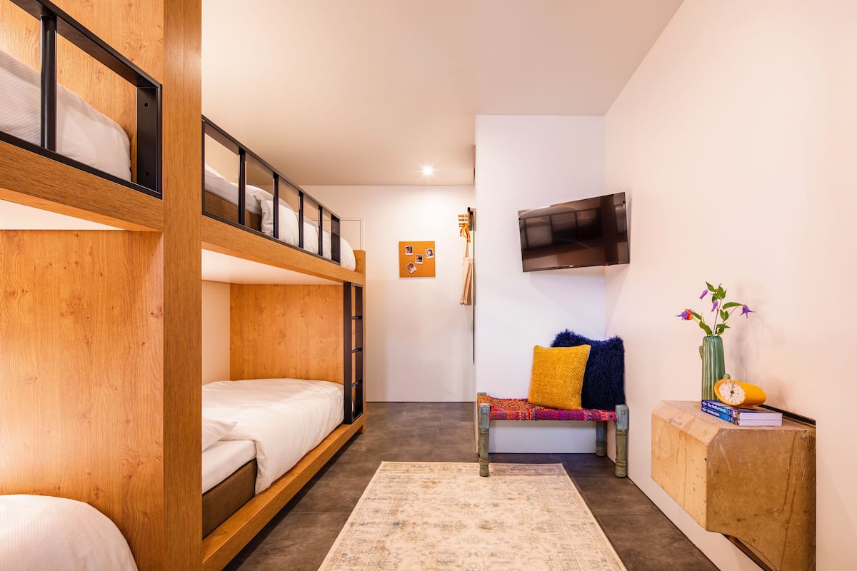 A room with bunk beds, a TV mounted on the wall, a small table with a vase and books, and a bench with cushions.