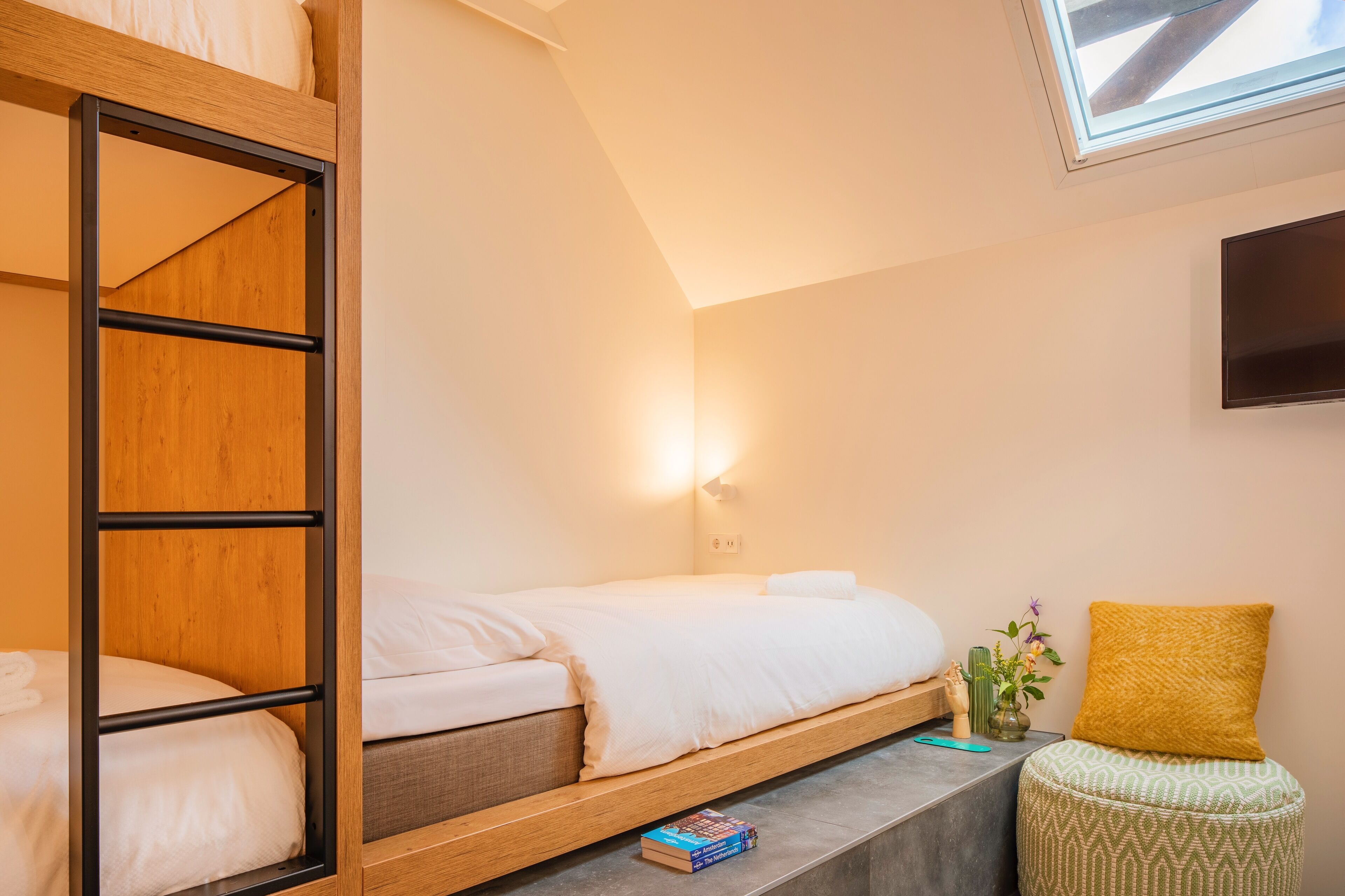 Una cama litera con estructura de madera y ropa de cama blanca, una mesita de noche con un libro, una pequeña planta en maceta y un cojín amarillo.