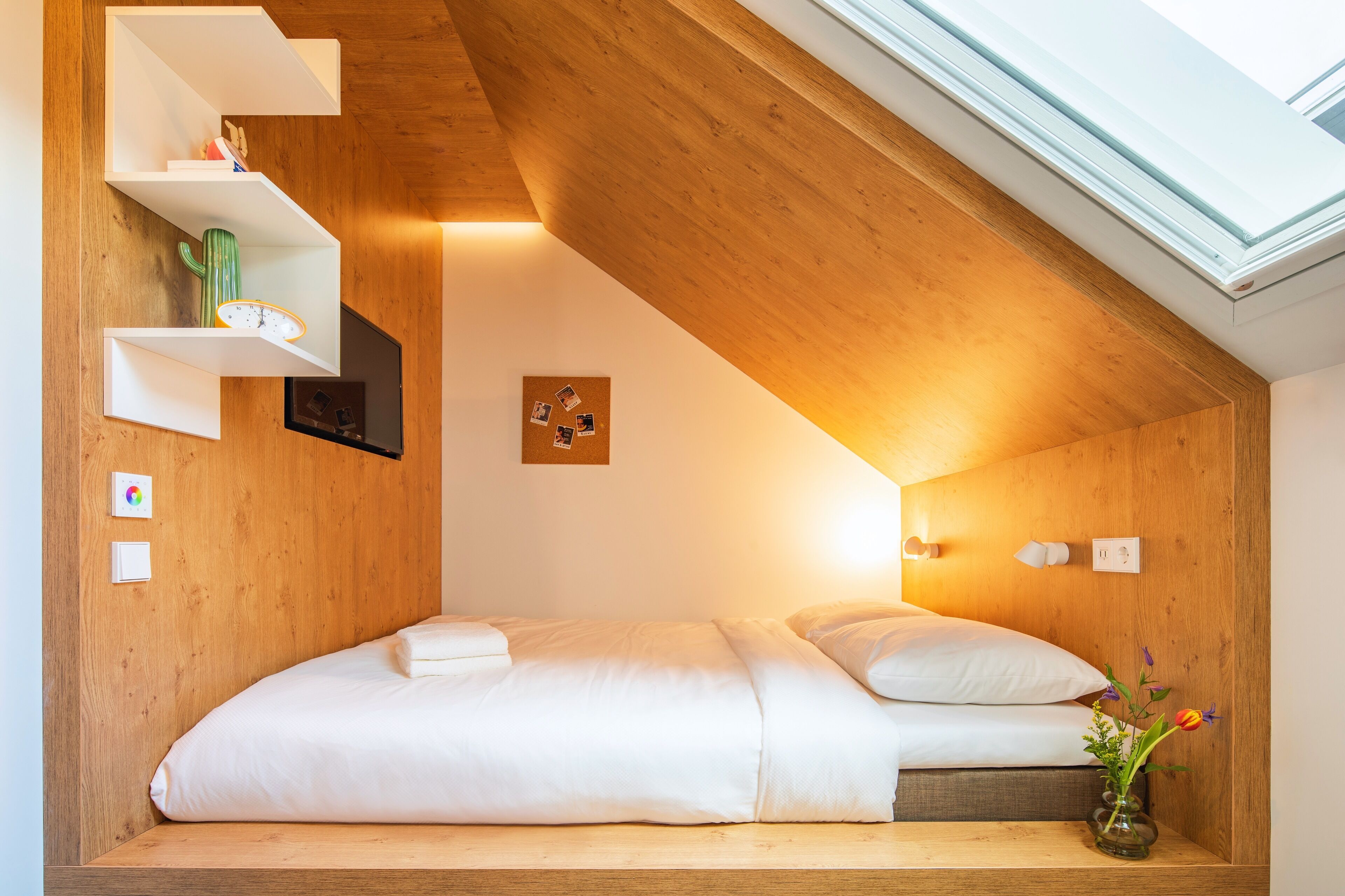 A cozy bedroom with a sloped ceiling, a bed with white bedding, a cactus on a shelf, and a vase with flowers.