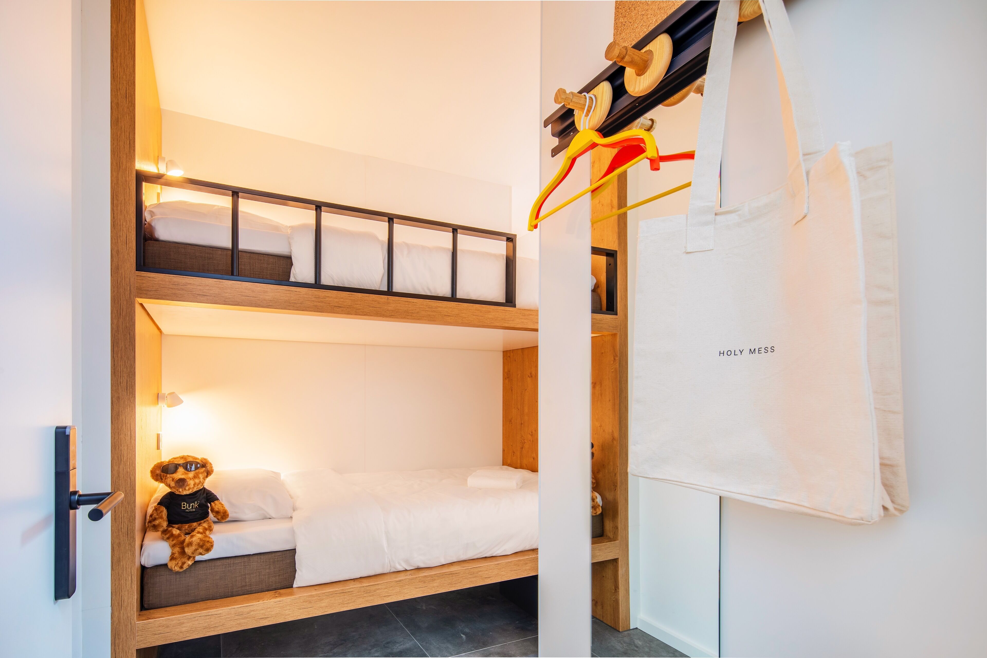 Habitació amb lliteres, un os de peluix al llit inferior, una tovallola blanca i una bossa amb la inscripció 'HOLY MESS' a la paret.