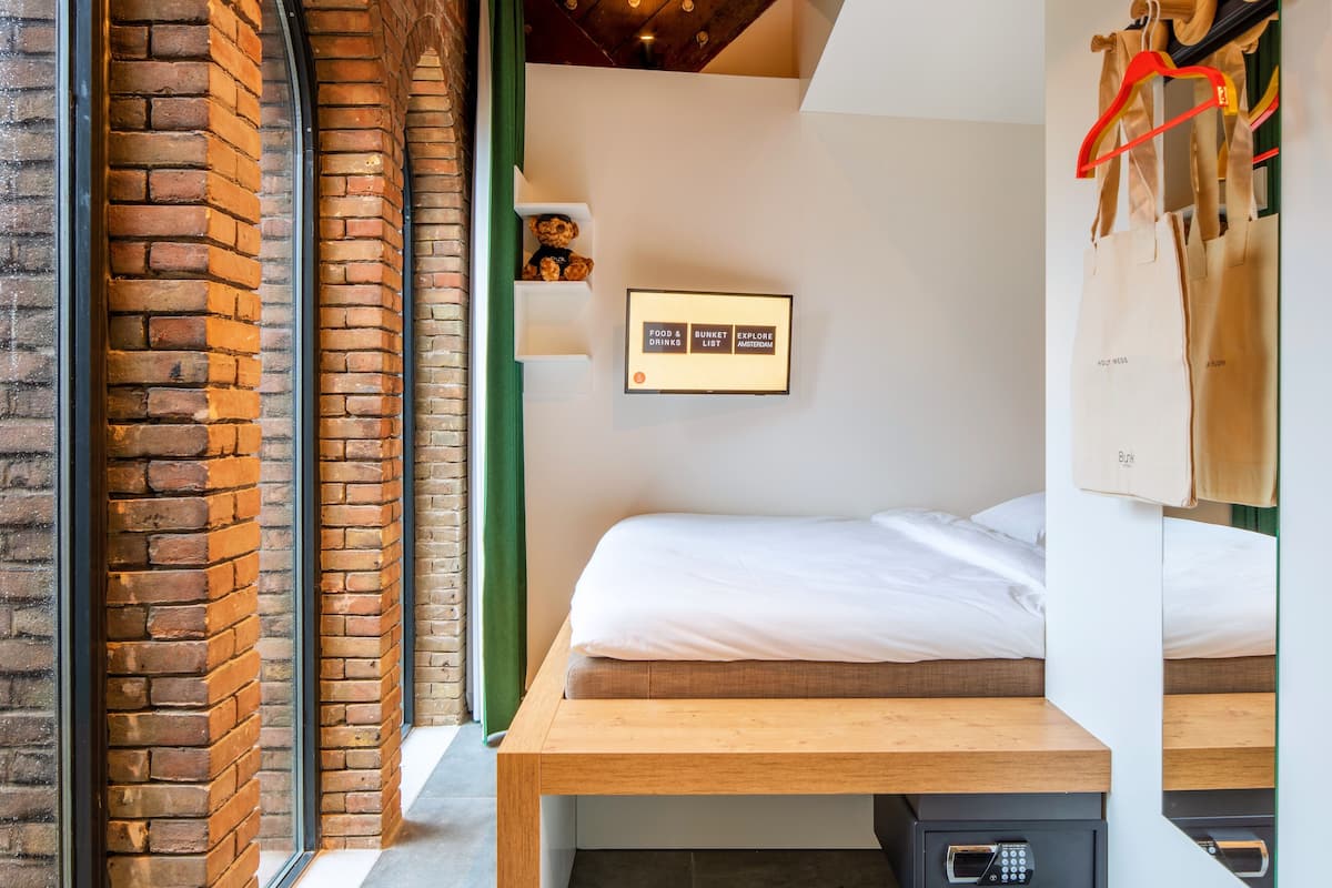 A room with a brick wall, a bed with white bedding, a wooden bench, a small shelf with snacks, and a sign with food and drink options.