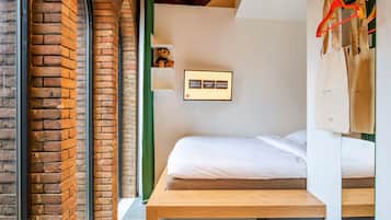 A room with a brick wall, a bed with white bedding, a wooden bench, a small shelf with snacks, and a sign with food and drink options.