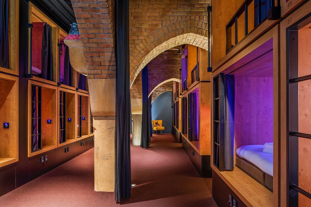 A hotel corridor with wooden bunk beds, a brick archway, and a purple-lit room in the distance.