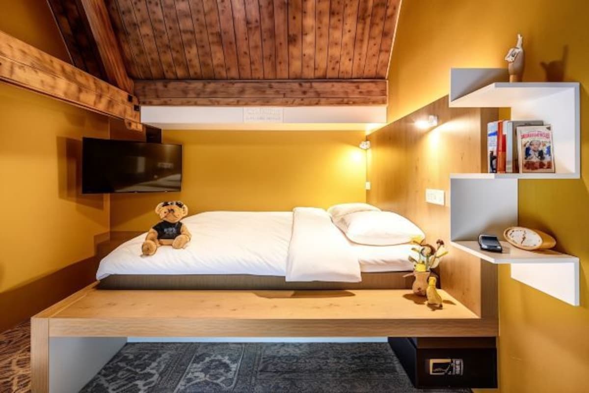 A cozy room with a bed, a teddy bear, a shelf with books, and a wall-mounted TV.