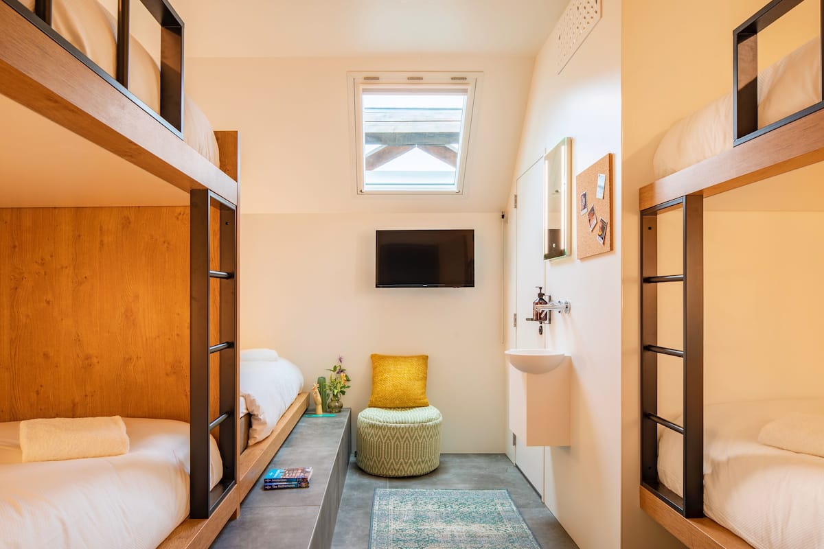 A compact hotel room with bunk beds, a small stool, a flat-screen TV, and a window with blinds.