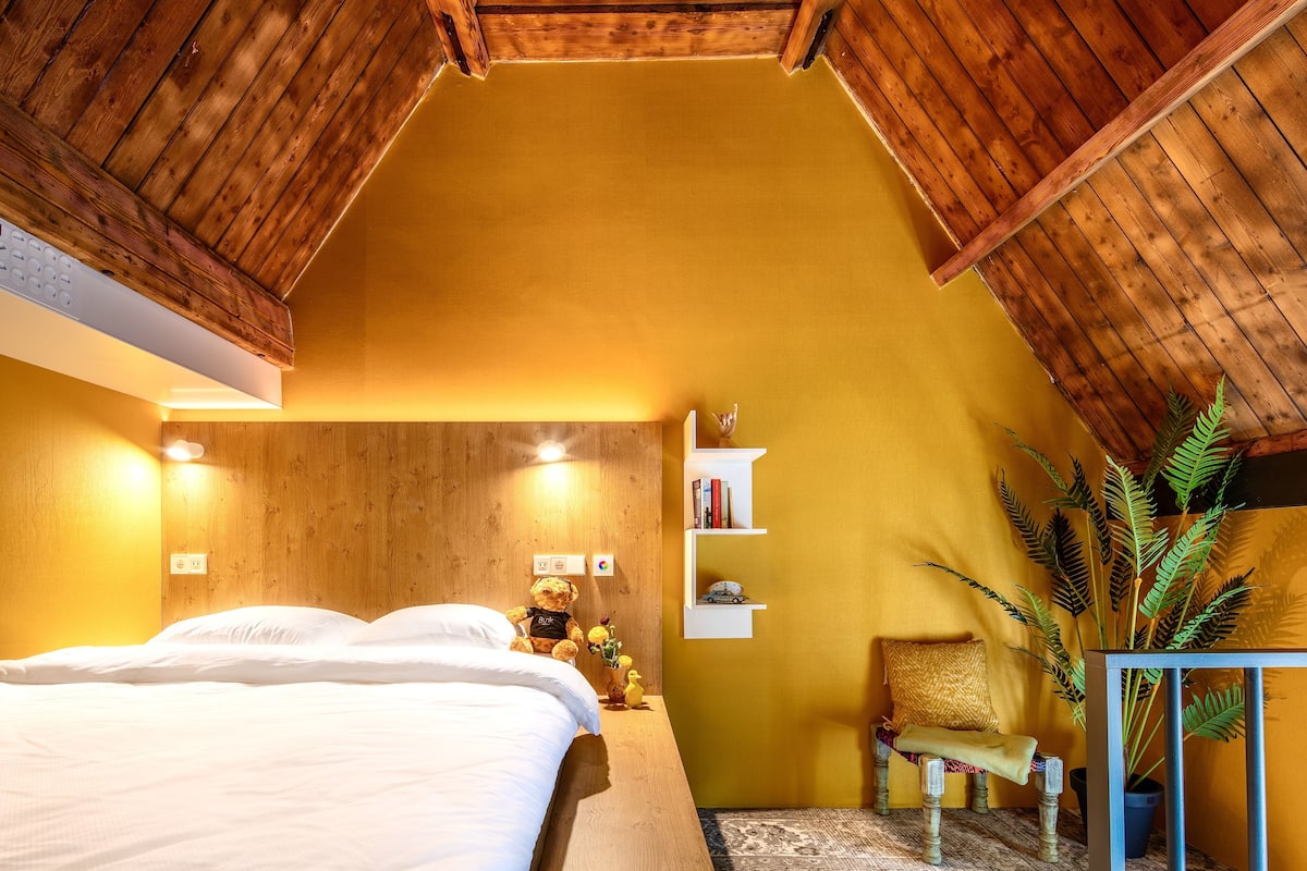 A cozy bedroom with a wooden ceiling, a bed, a nightstand, a chair, a shelf with books and a plant, and a small table with a plant.