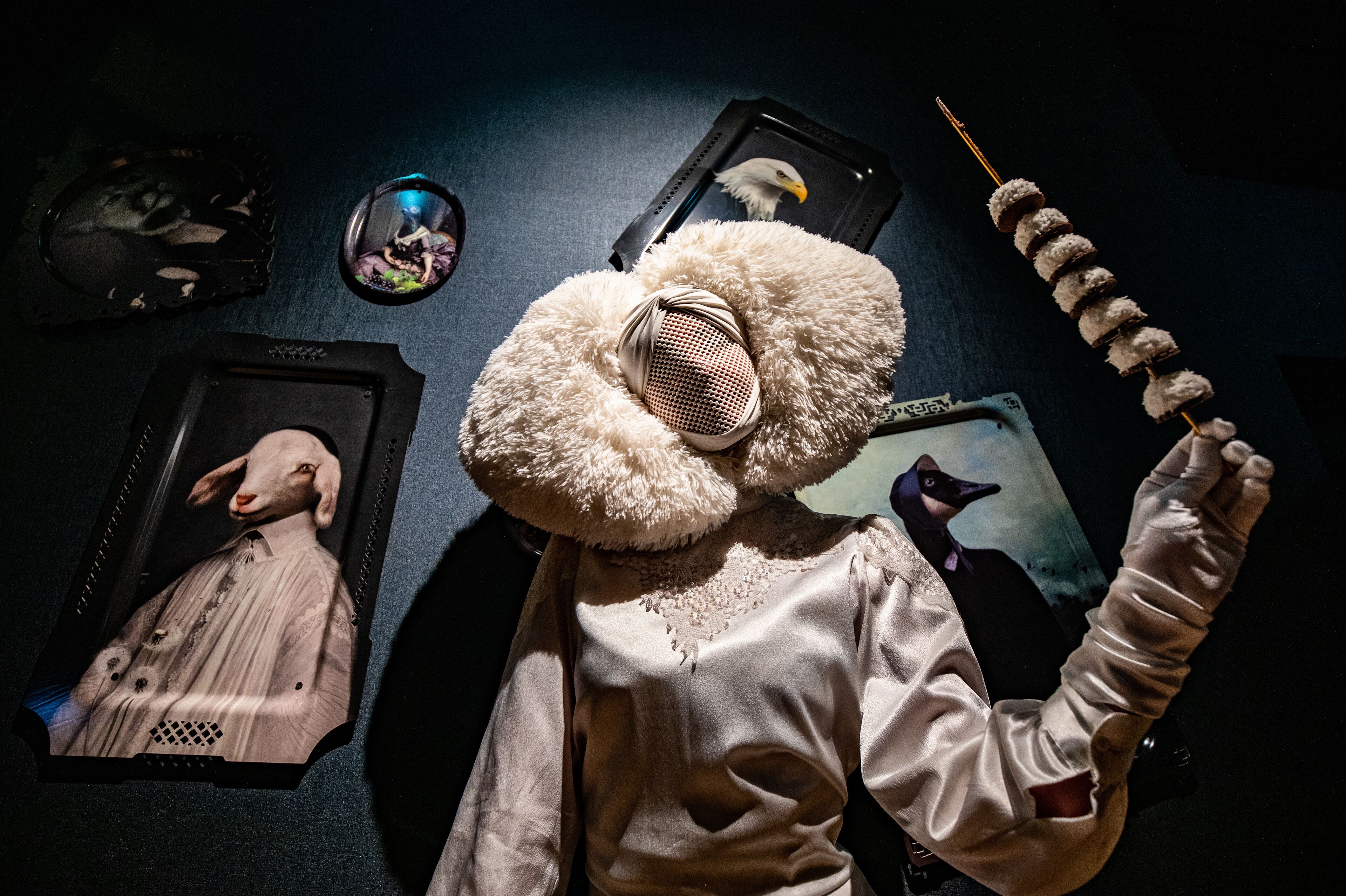 A mannequin with a large headpiece and a long stick, surrounded by framed pictures on the wall.