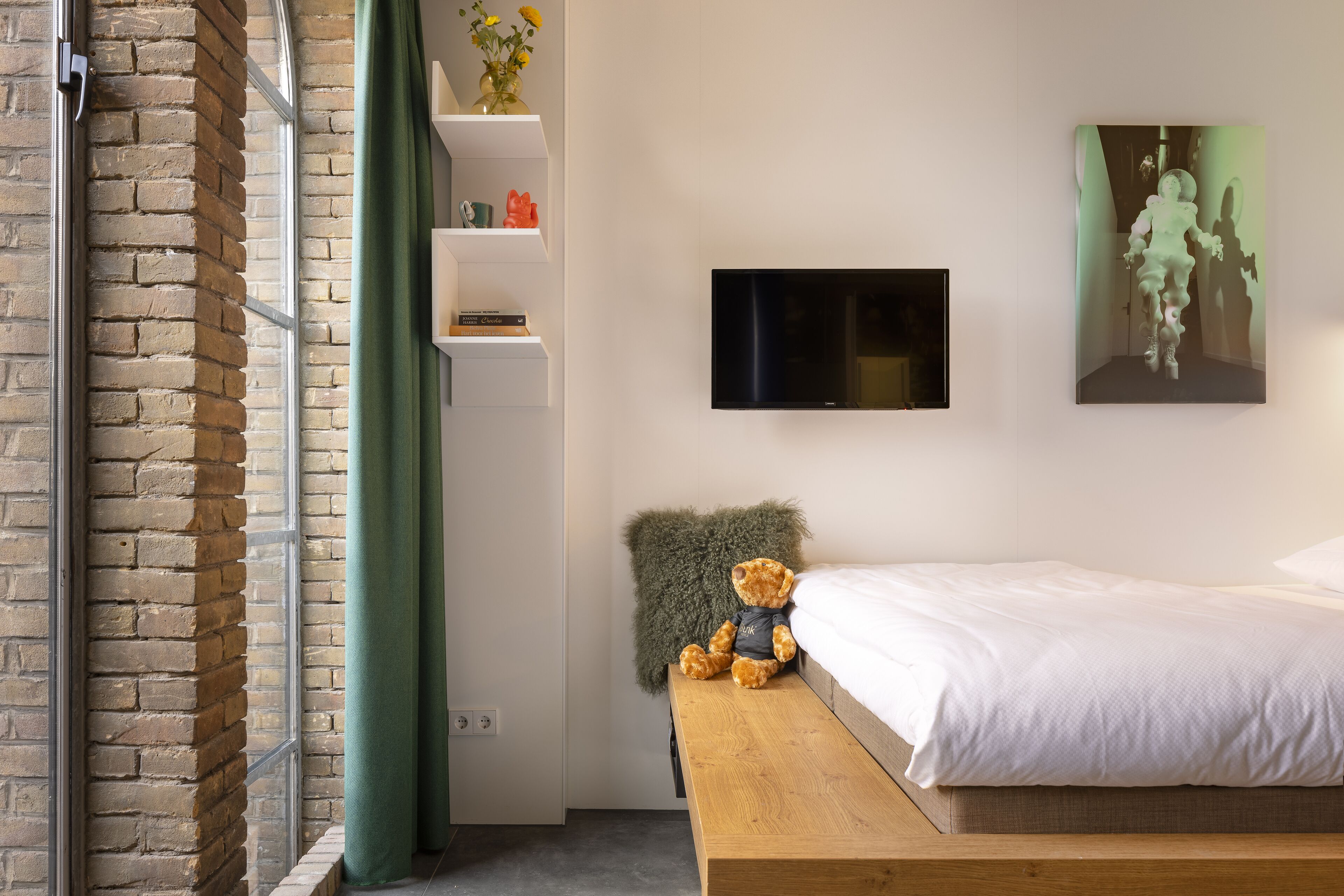 Un dormitorio con pared de ladrillo, cortina verde, banco de madera, cama, televisor y estante con objetos decorativos.