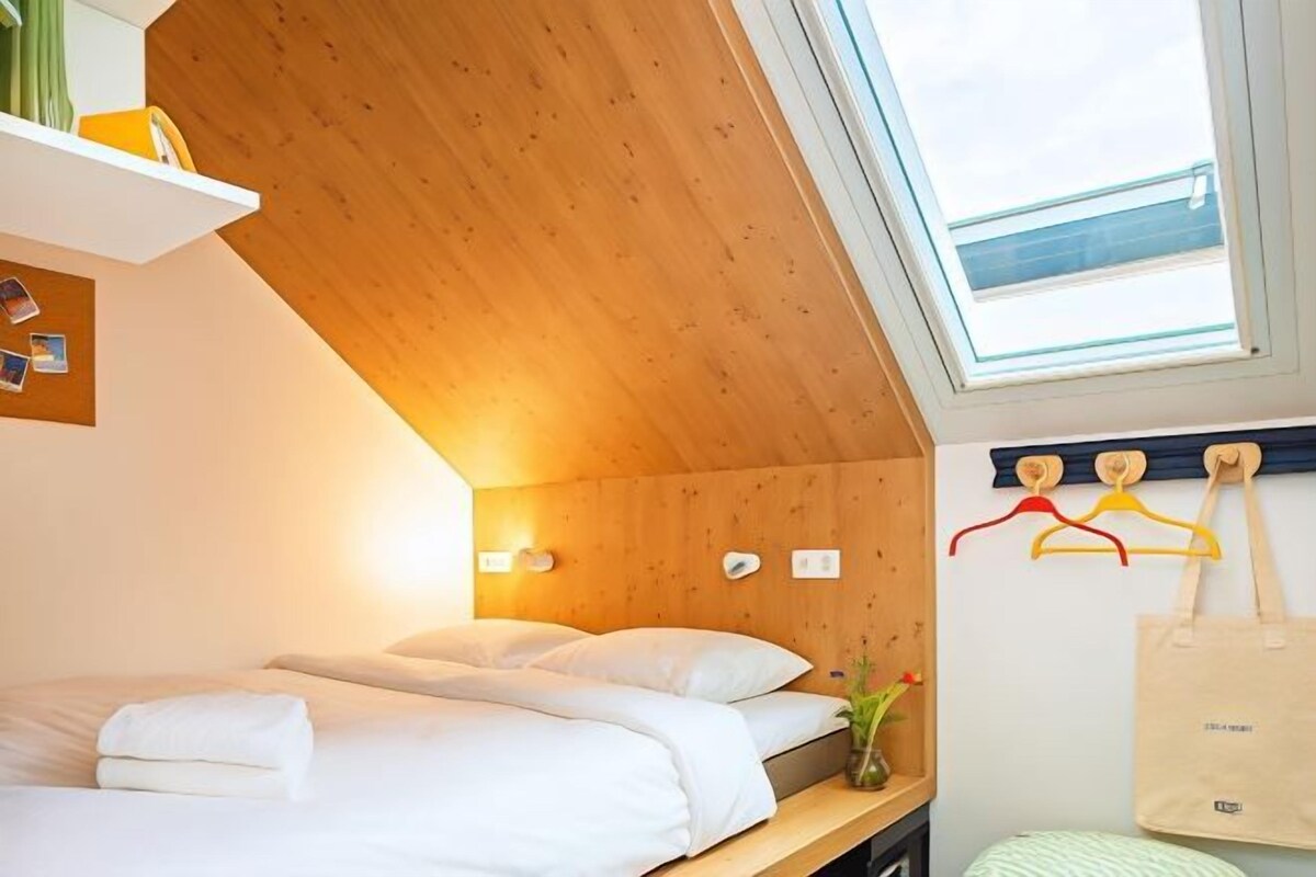 Un dormitorio acogedor con techo de madera, tragaluz, cama con ropa blanca, mesita de noche con un florero y perchero con ganchos.