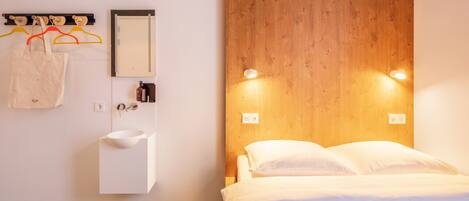 Une chambre d’hôtel moderne avec une tête de lit en bois, une table de chevet, un lavabo blanc et un miroir.