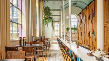 A spacious dining area with long tables and chairs, large windows, and hanging plants.
