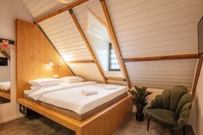 A bedroom with a sloped ceiling, wooden bed frame, white bedding, a green armchair, and a potted plant.