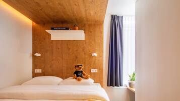 Un dormitorio con techo de madera, una cama con ropa de cama blanca, un oso de peluche en la cama, un estante con un libro y una planta, y una ventana con cortinas.