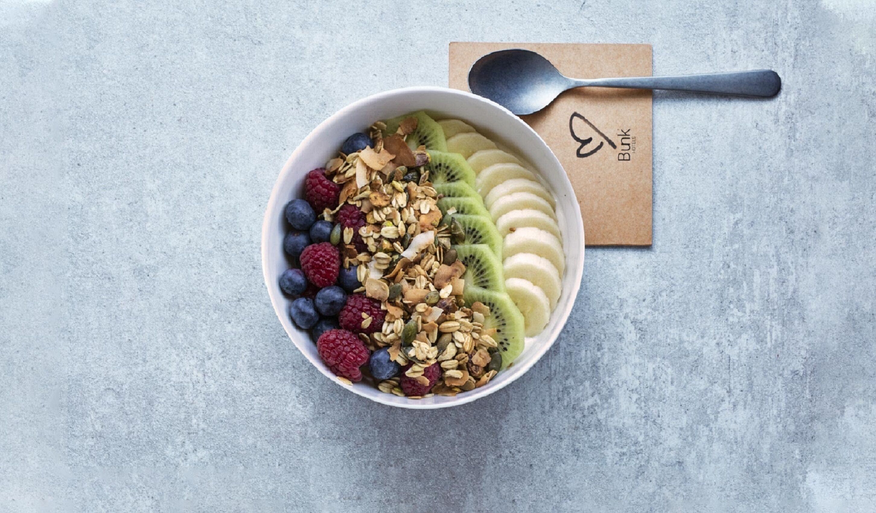A bowl of fruit and granola on a textured surface with a spoon and a napkin with a logo.