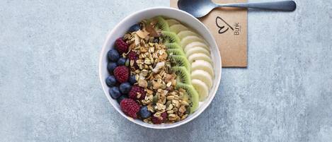 A bowl of fruit and granola on a textured surface with a spoon and a napkin with a logo.