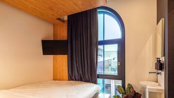 Une chambre d’hôtel moderne avec un lit, une grande fenêtre avec des rideaux, un lavabo blanc et un miroir.