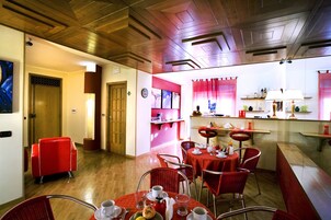 Uma área de refeições de hotel com cadeiras e mesas vermelhas, piso de madeira e uma cozinha ao fundo.