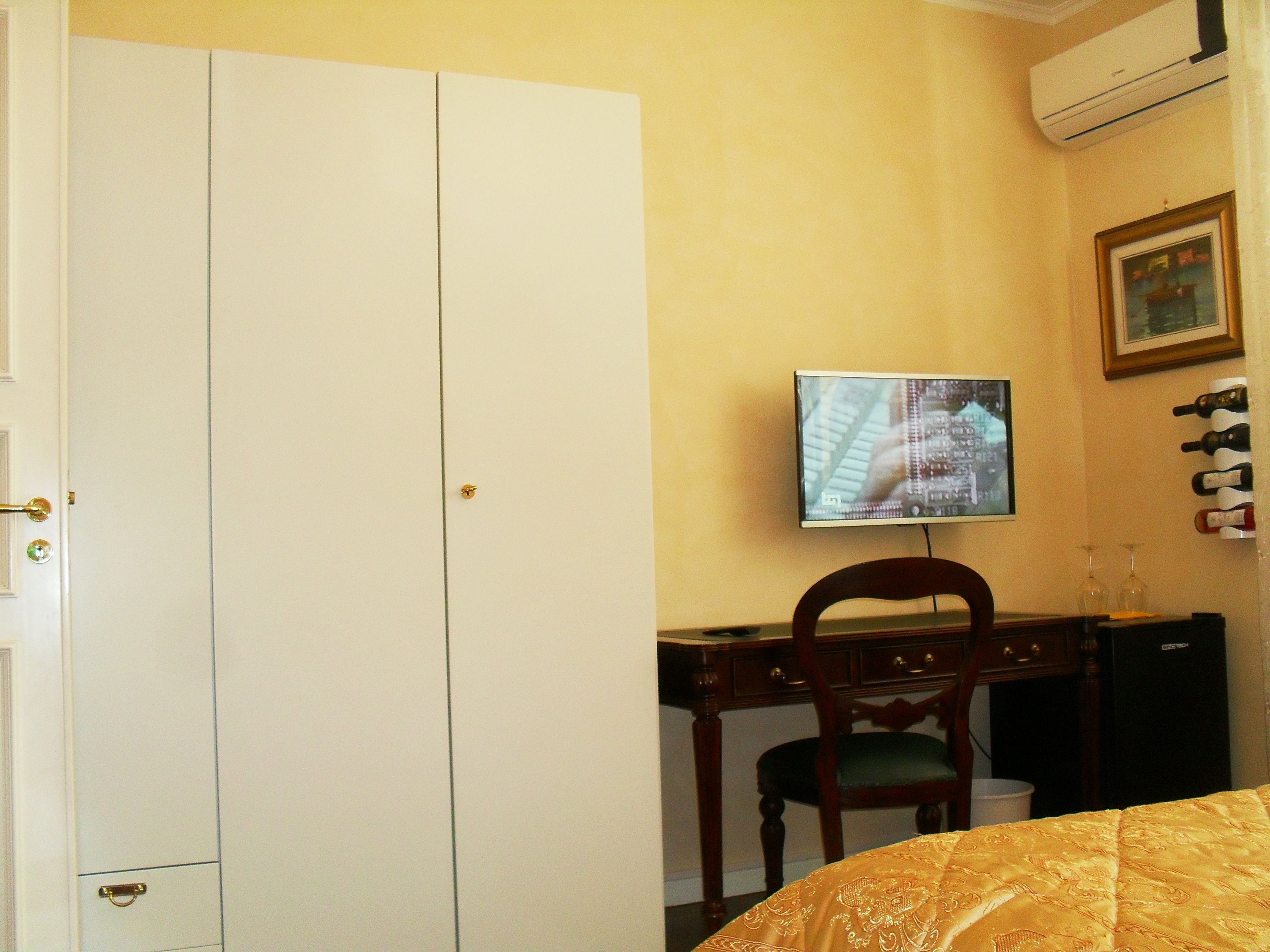 Habitación de hotel con cama, escritorio con silla, televisor y una estantería para vinos en la pared.