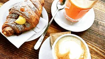 Un croissant saupoudré de sucre glace et garni d’une tranche de citron, sur une assiette, un verre de jus d’orange et une tasse de café décorée d’un motif en latte, le tout posé sur une table en bois.