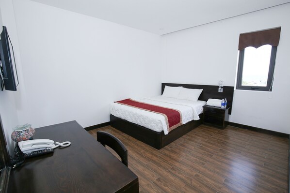 Hotelkamer met een bed, nachtkastje met een lamp, bureau met telefoon en raam met bruin gordijn.