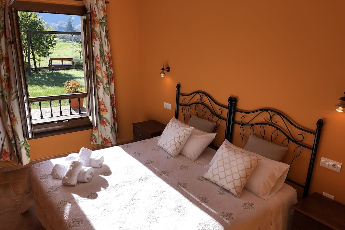 A bedroom with a bed, pillows, a nightstand, a window with curtains, and a view of a garden.