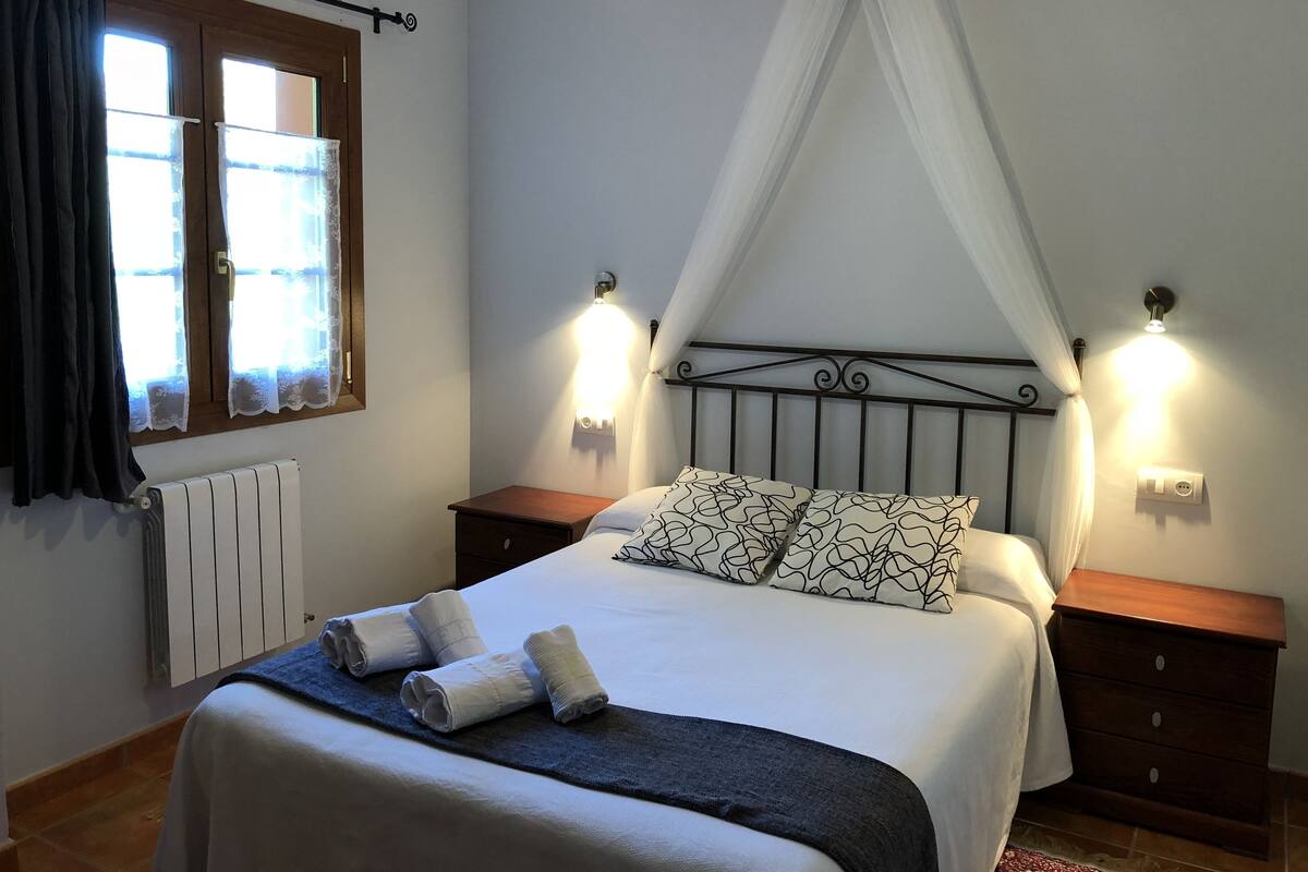 A neatly made bed with a canopy, bedside tables, a radiator, and a window with curtains.