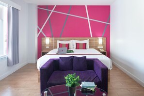 Una habitación de hotel moderna con una cama grande, un sofá púrpura, una mesa de centro de cristal y una planta en maceta.