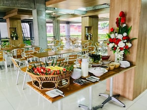 A dining area with tables set for a meal, featuring a central fruit basket and a floral arrangement. - Eco Inn Lite Ubon Ratchathani (Ubon Ratchathani)