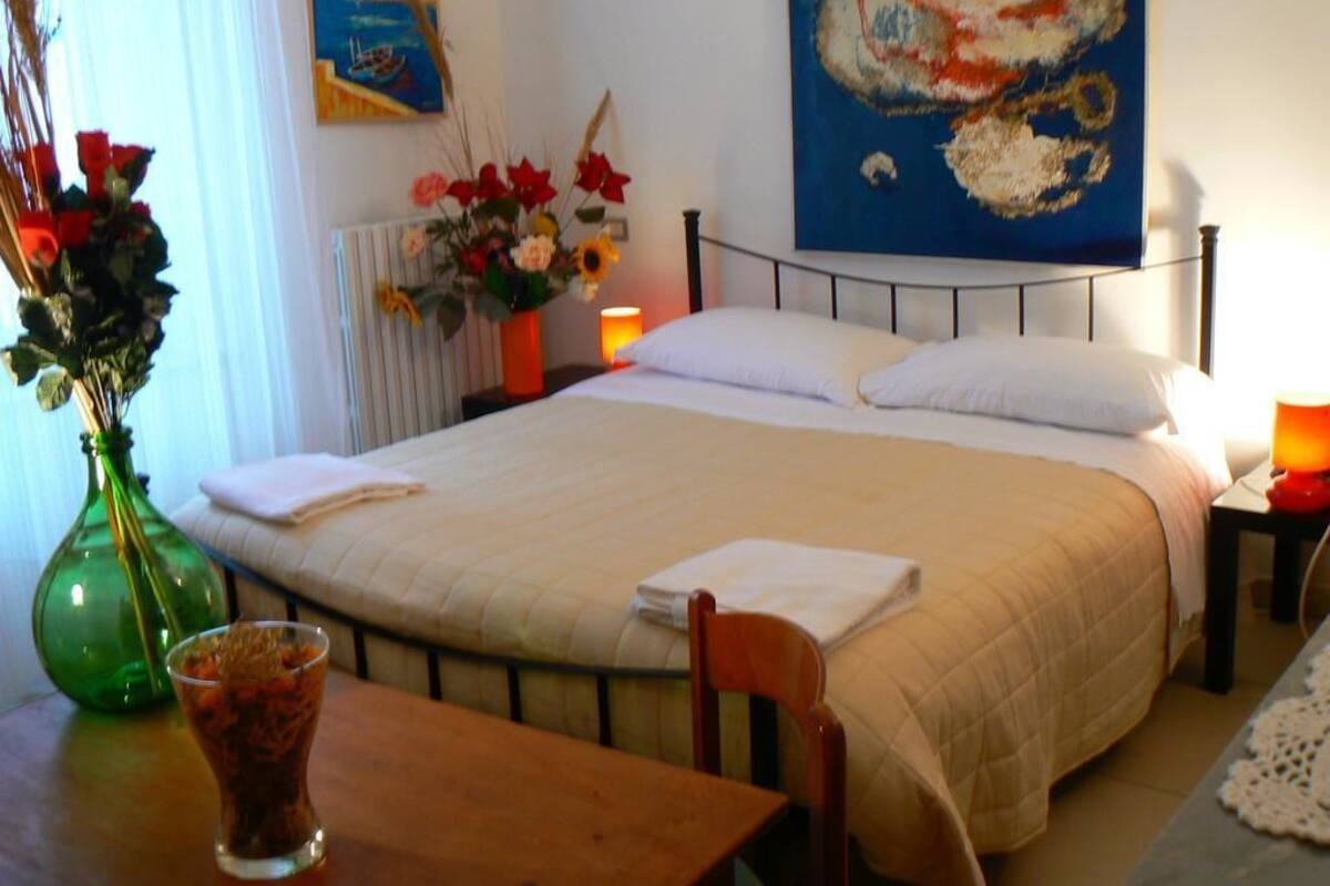 Ein ordentlich bezogenes Bett mit Stecken, eine Vase mit Blumen, ein Tisch mit einem Glas und ein Gemälde an der Wand.