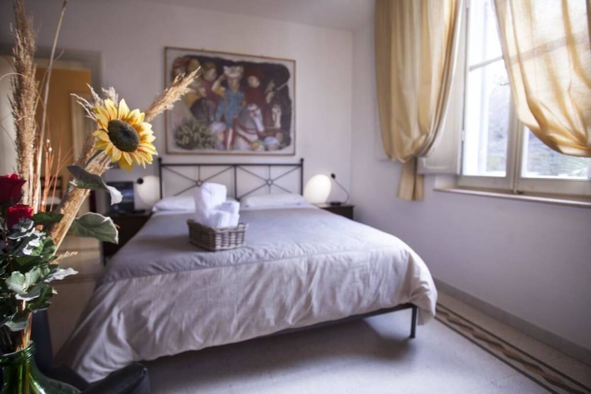 Ein Schlafzimmer mit Bett, Nachttisch, Lampe, einem Bild an der Wand und einem Fenster mit Vorhängen.