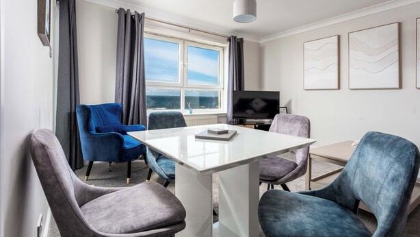 A modern room with a white table, four chairs, a flat-screen TV, and a large window with curtains. - Carrick Retreat - Donnini Apartments (Ayr)