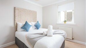 A neatly made bed with a white comforter, two blue pillows, and a rolled white towel. A gray throw blanket is draped over the bed. - Carrick Retreat - Donnini Apartments (Ayr)