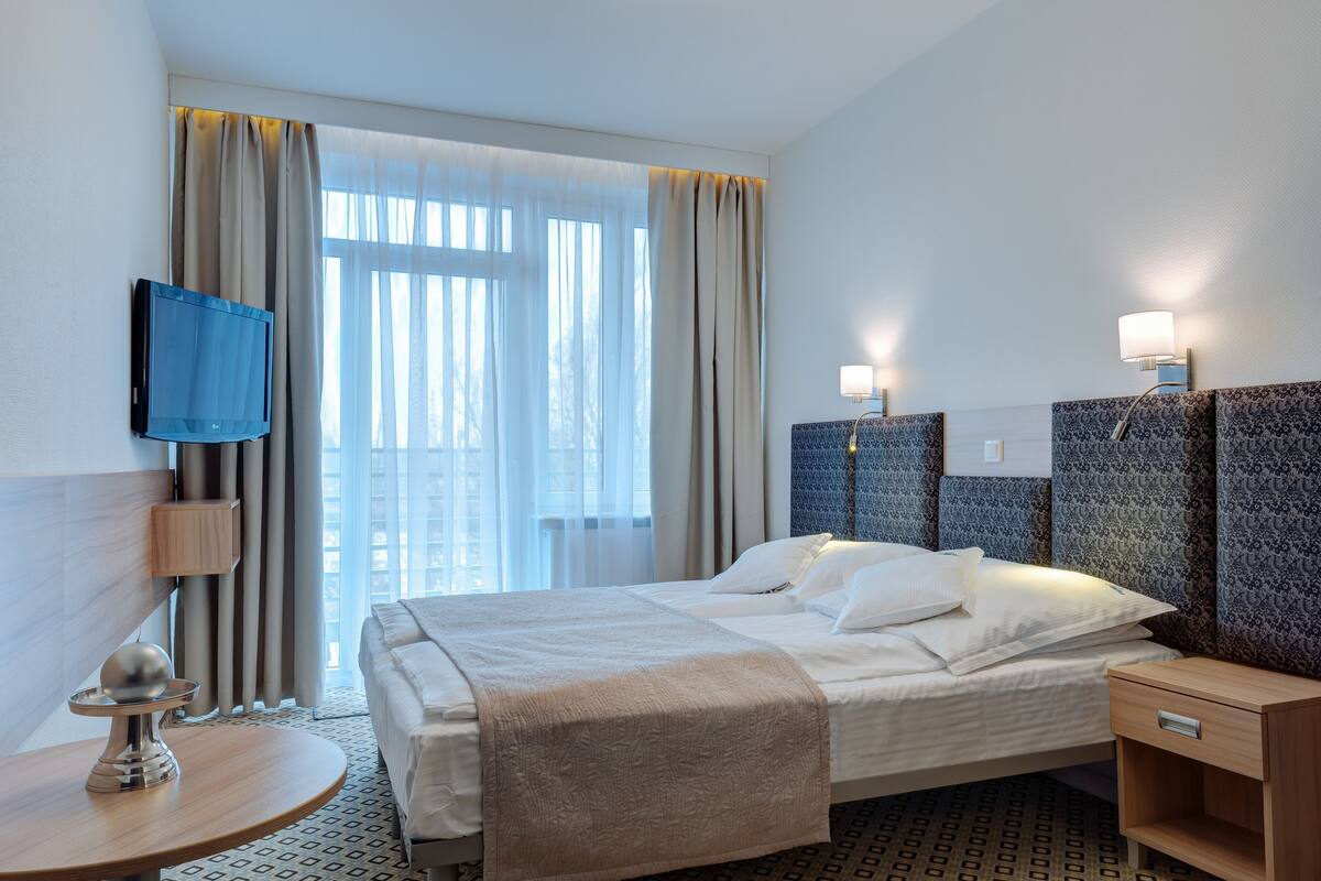 Een hotelkamer met een bed, een televisie, een nachtkastje, een lamp en een raam met gordijnen.