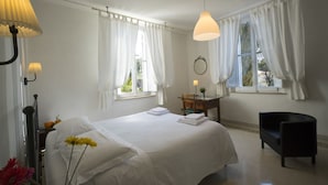 Een slaapkamer met een bed, twee ramen met gordijnen, een nachtkastje met een lamp, een stoel en een spiegel aan de muur.