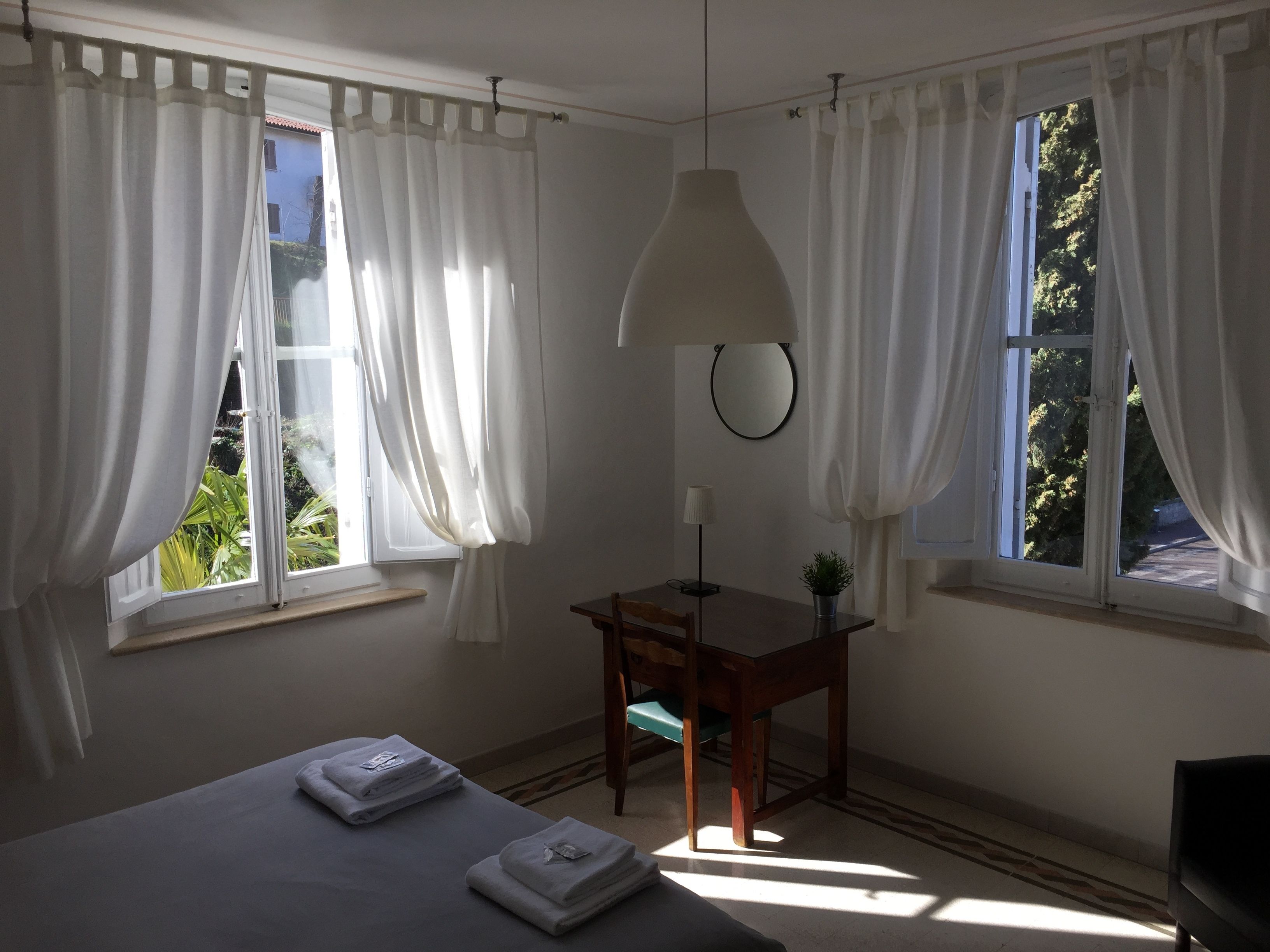 A room with two windows, a bed with towels, a desk with a lamp, and a chair.