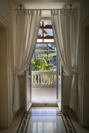 An open doorway with white curtains, a tiled floor, and a view of a balcony with a railing and greenery outside.