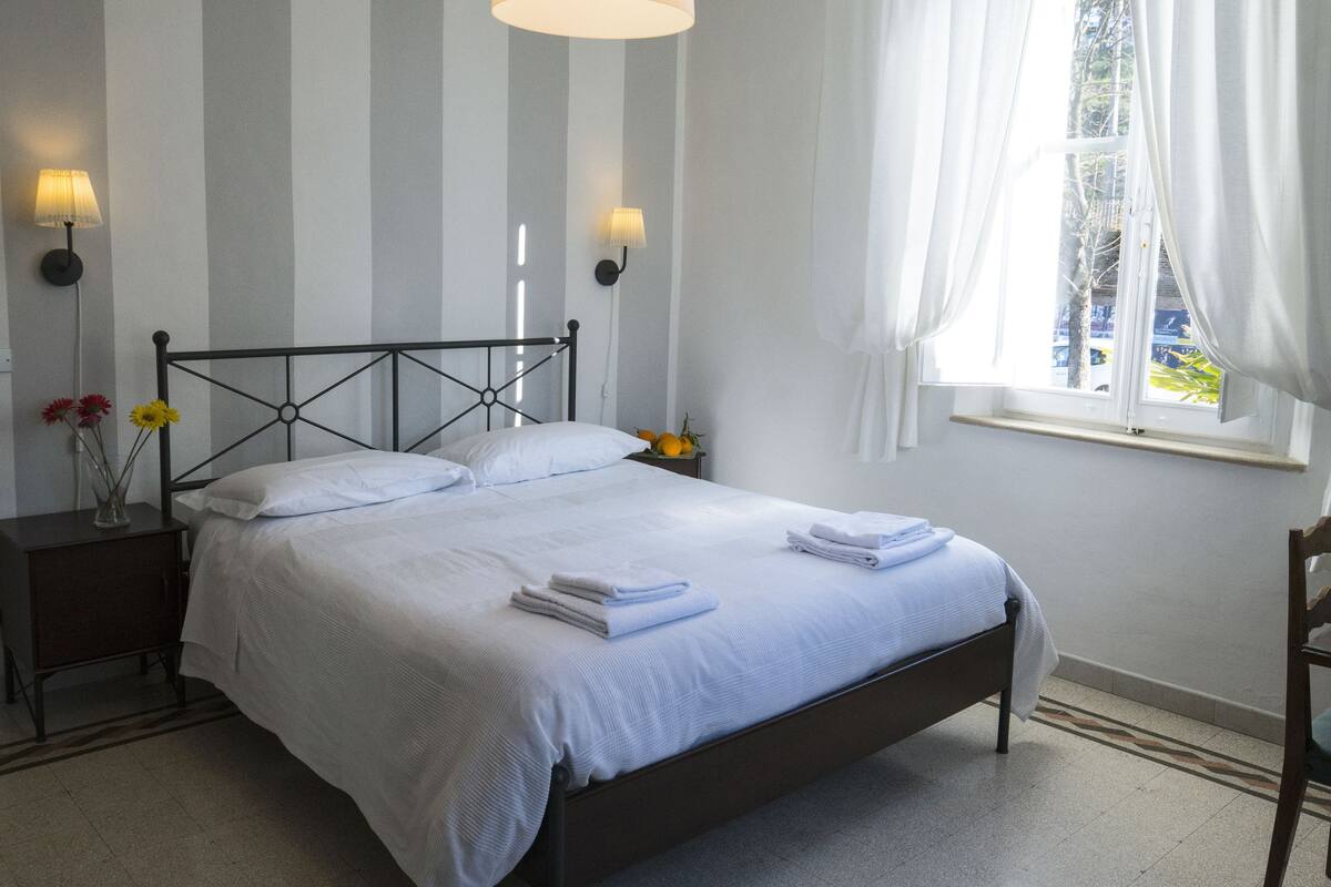 A neatly made bed with white linens, a bedside table with a lamp, and a window with curtains.