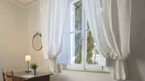 Een kamer met een houten bureau, een stoel, een zwarte fauteuil, een raam met witte gordijnen en een kleine potplant.