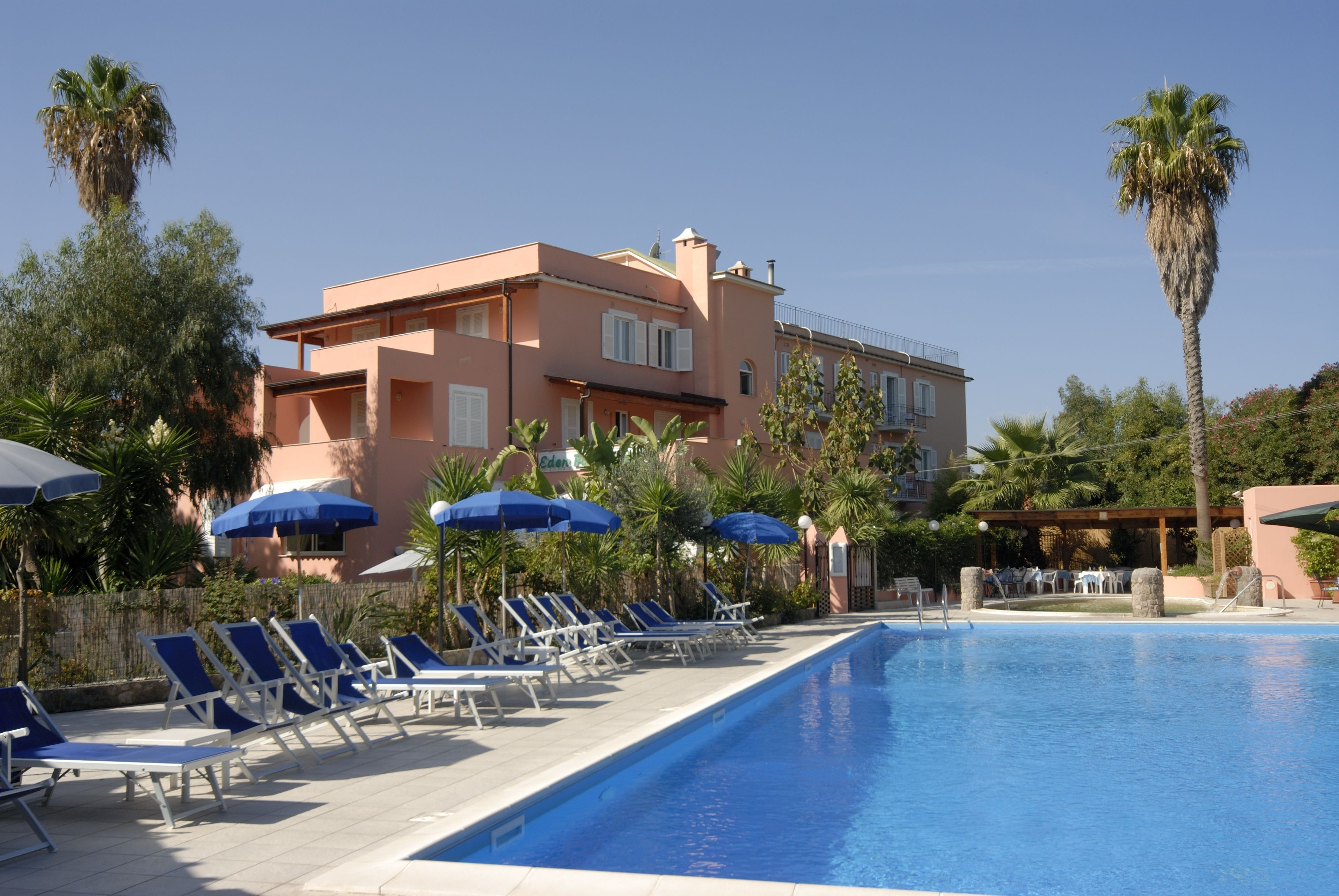 Ein Poolbereich mit Liegestühlen und Sonnenschirmen, ein Gebäude mit rosa Fassade und Palmen.