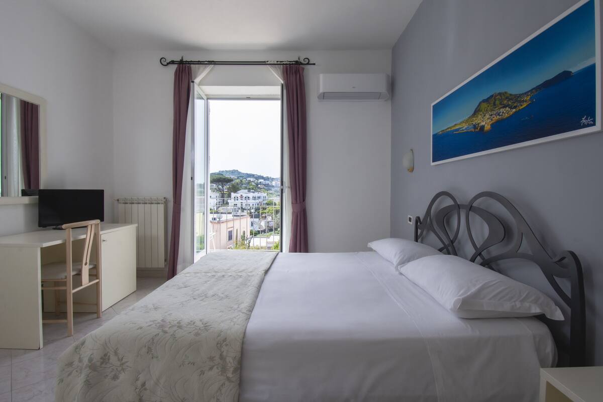 A hotel room with a bed, a desk with a chair, a television, and a window with curtains.