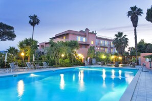 A swimming pool with lounge chairs and palm trees, surrounded by a building with balconies and warm lighting. - Eden Park (Forio)
