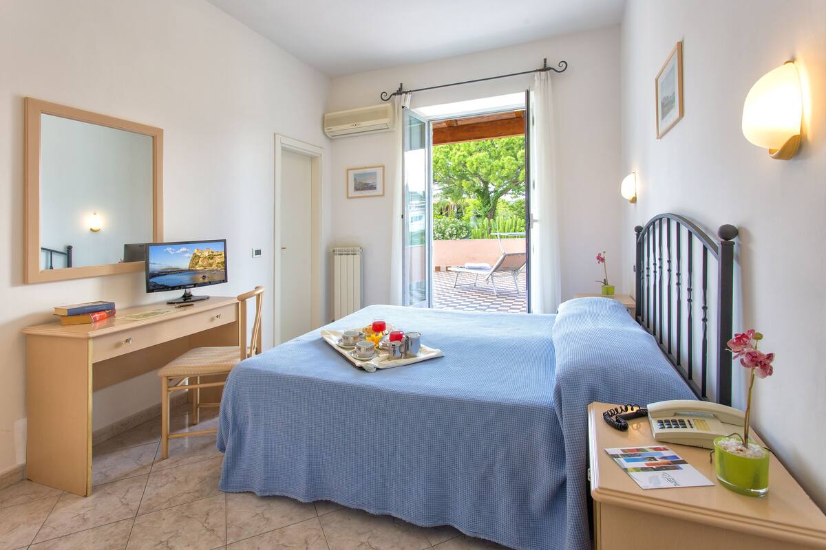 A hotel room with a bed, a desk with a computer, a chair, a small table with a tray of breakfast items, a bedside table with a phone and a vase with flowers, and a window with a view of greenery.