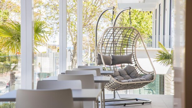 A modern outdoor seating area with a wicker swing chair and cushioned chairs, large windows, and potted plants. - Hotel Amalfi riccione (Riccione)