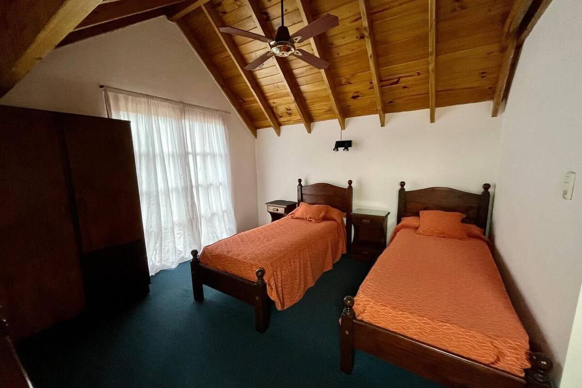 Quarto com duas camas de madeira, um ventilador de teto e uma janela com cortinas.