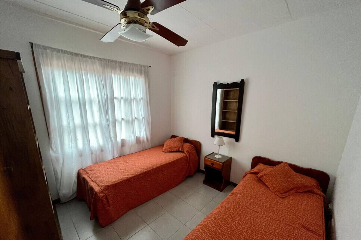 Quarto com duas camas, um ventilador de teto, uma janela com cortinas, uma mesinha de cabeceira com abajur e um espelho na parede.
