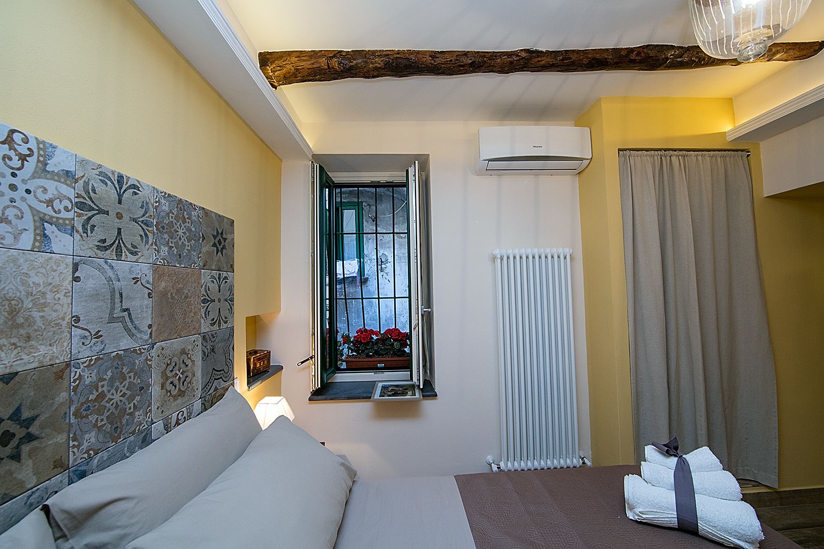 Un dormitorio con cama, ventana con flores, radiador y cortina.