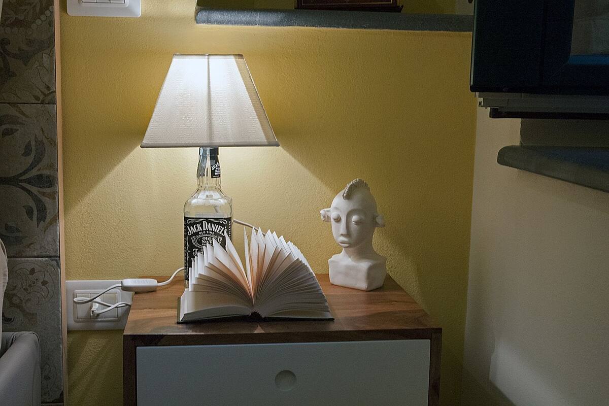 Una mesita de noche con una lámpara, un libro abierto, una botella de whisky Jack Daniel's y un busto.