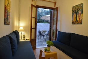 Ein gemütliches Wohnzimmer mit zwei dunkelblauen Sofas, einem kleinen Holztisch mit einer Pflanze und einer Lampe. Eine offene Tür führt auf eine Terrasse mit Tisch und Stühlen.
