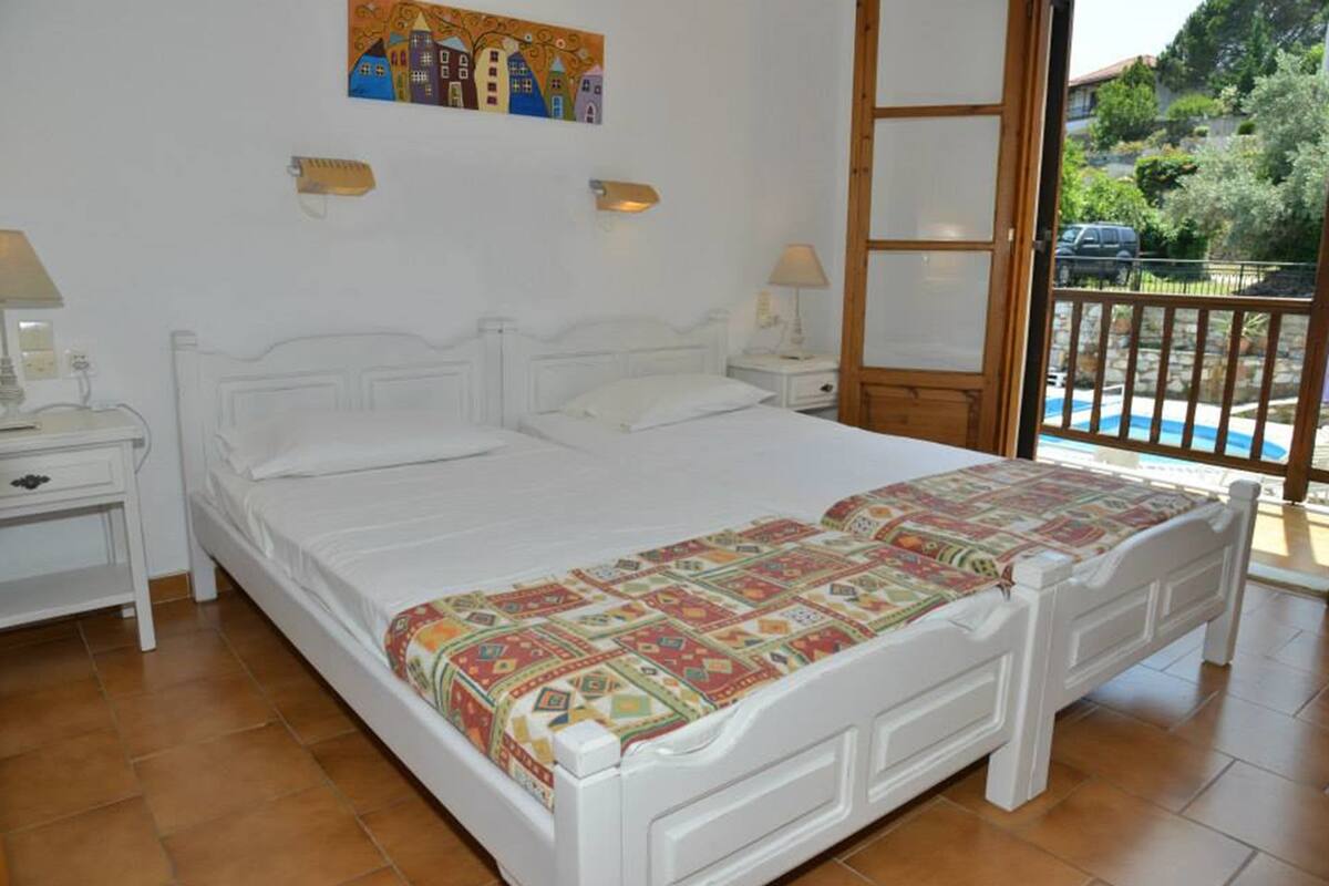 Un dormitorio con una cama grande, cabecera blanca, dos mesitas de noche con lámparas, una colcha colorida y vista a una piscina a través de una puerta abierta.