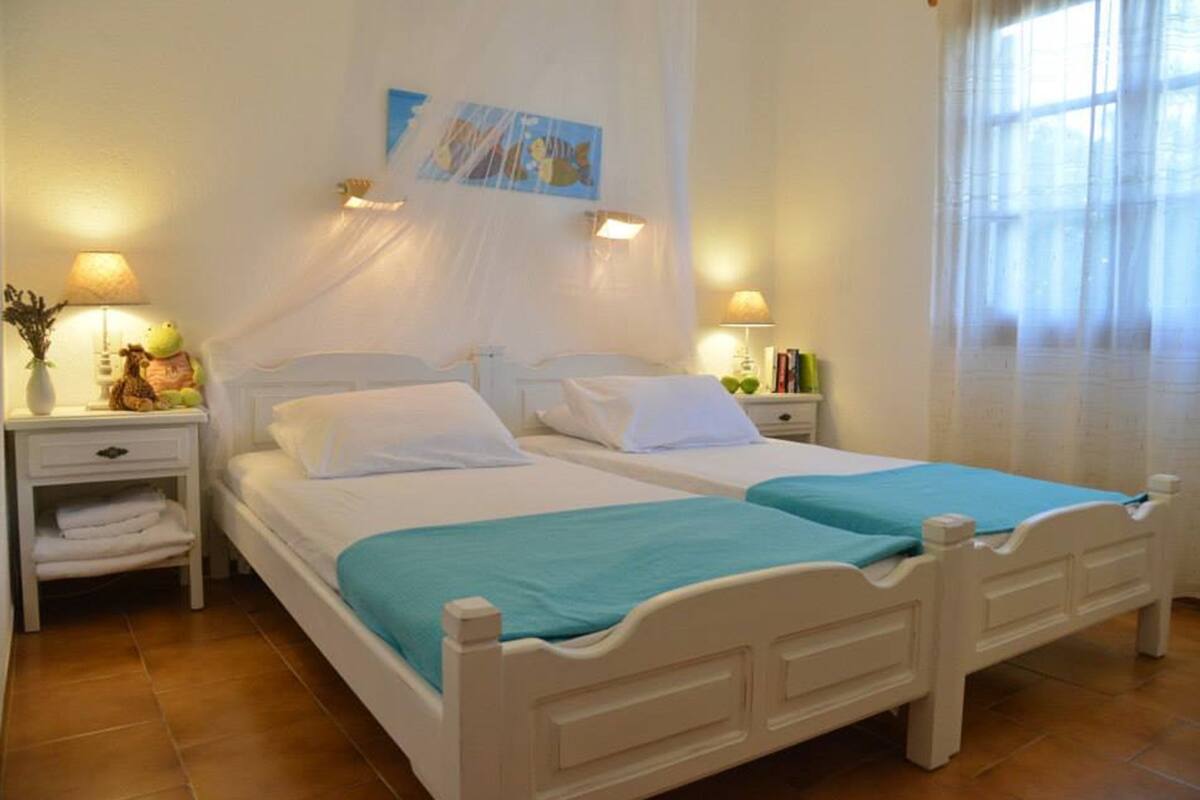 Una cama doble con ropa de cama blanca y un plaid verde azulado, dos mesitas de noche con lámparas, una obra de arte colgada en la pared y una ventana con cortinas traslúcidas.
